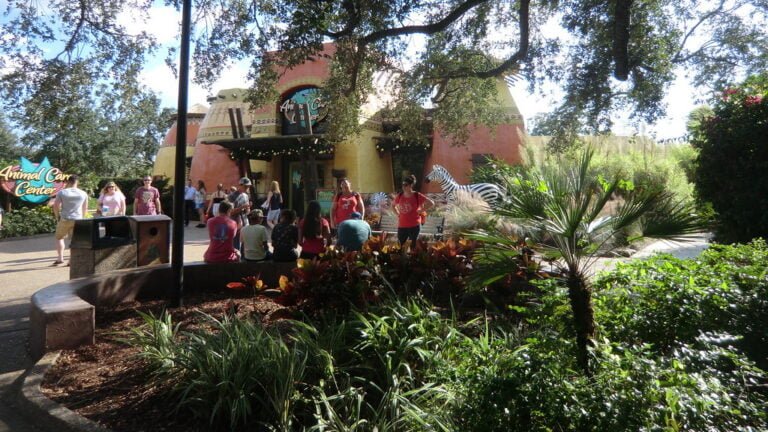 Visitors gather outside a colorful building with a sign reading "Animal Care Center," surrounded by lush greenery and a partial view of a zebra statue to the right.