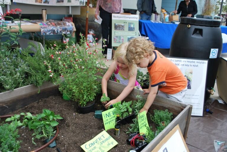 Two young children are gardening together in a raised flower bed with various plants at an outdoor educational event, with informational signs and posters about composting and water conservation visible in the background.