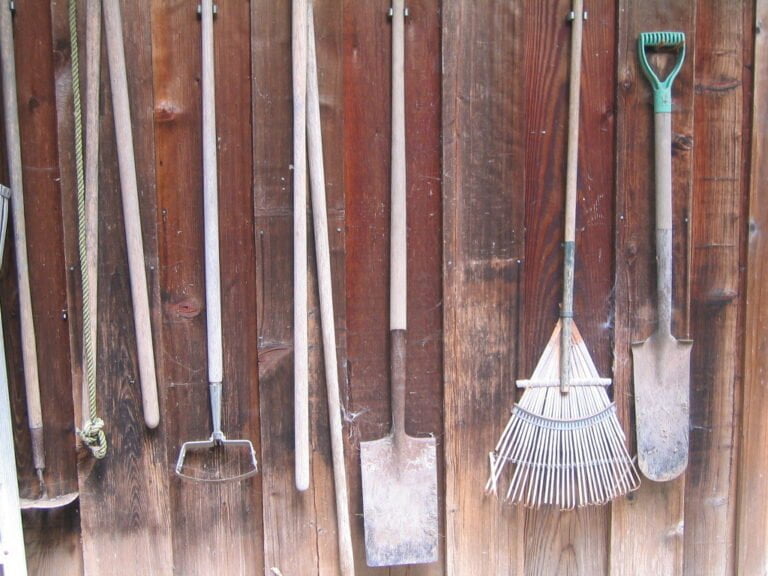 Assorted gardening tools, including rakes and shovels, hanging against a wooden slat wall.
