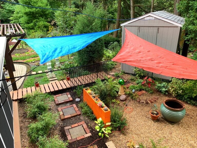 A colorful backyard garden with red and blue fabrics hanging to dry, a curved walking path, a garden shed, and various plants and garden decorations.