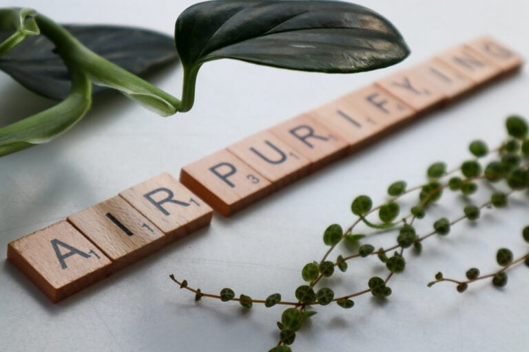 Scrabble tiles arranged on a white surface spelling "AIR PURIFYING" with parts of green plants on both sides of the words.