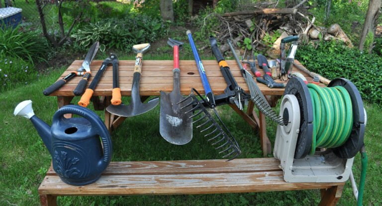 An array of gardening tools laid out on a wooden picnic table, including a black watering can, various trowels, a shovel, a fork, secateurs, a rake, and a hose reel, set against a background of a green garden.