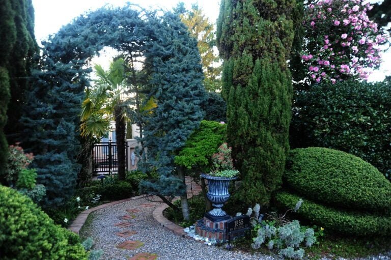 A serene garden pathway with neatly trimmed bushes, a decorative urn planter, and an archway formed by pruned trees, with a burst of pink flowers in the background.