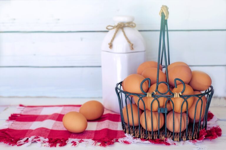 A wire basket filled with brown eggs on a red and white checkered cloth, with a white ceramic milk bottle in the background against a white wooden backdrop.