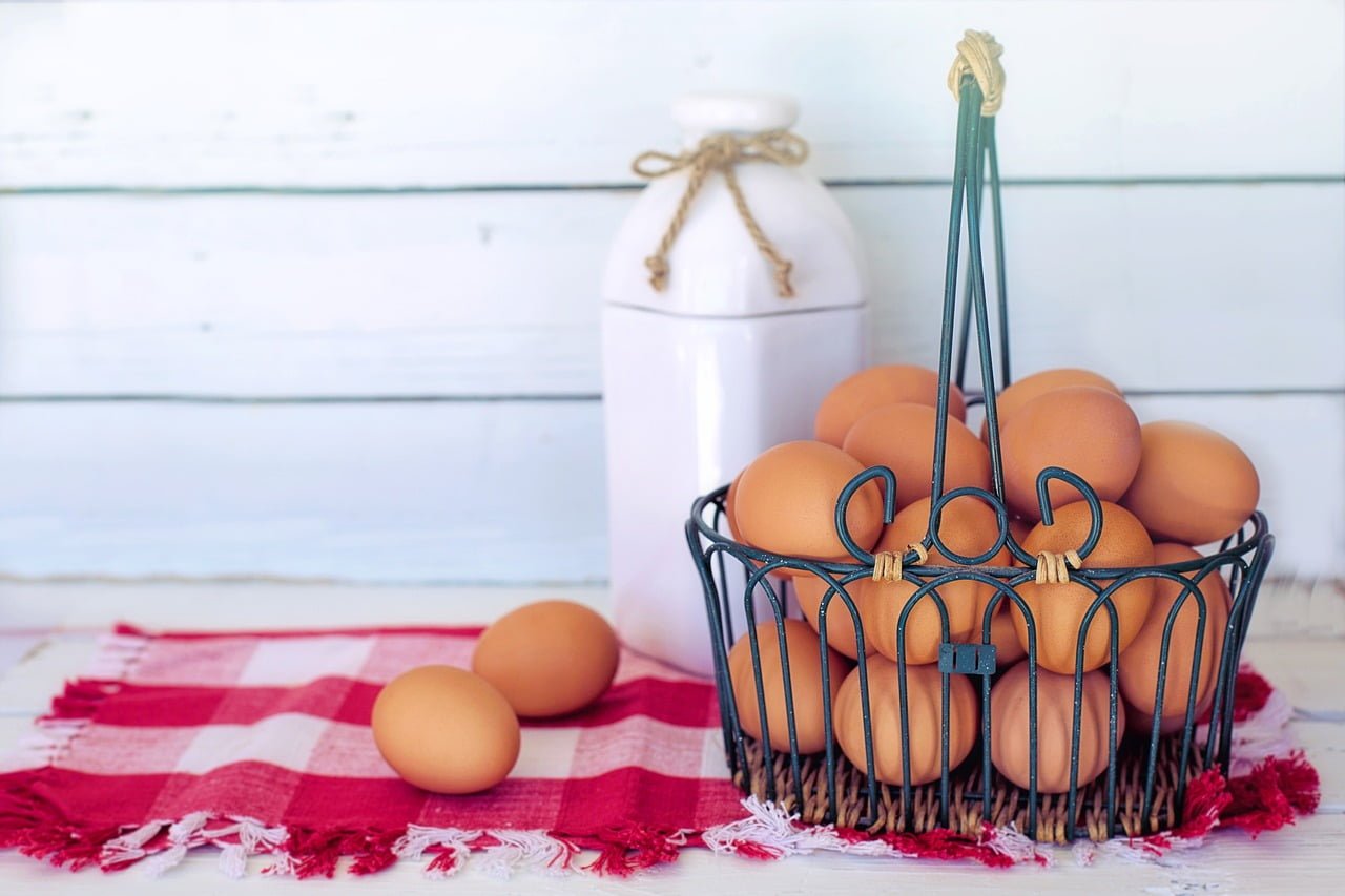 A wire basket filled with brown eggs on a red and white checkered cloth, with a white ceramic milk bottle in the background against a white wooden backdrop.