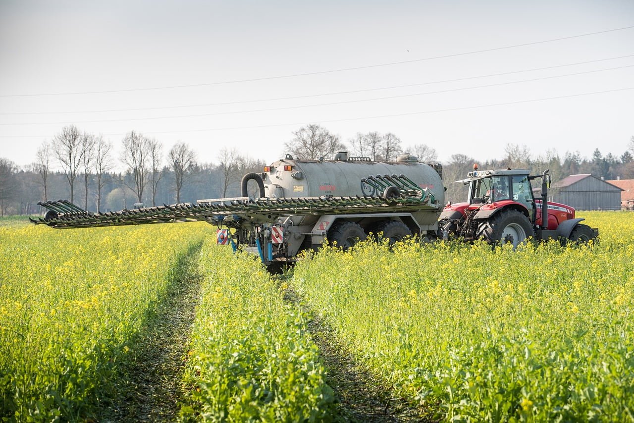 A red tractor pulling a large tank sprayer through a field of yellow flowering crops under a clear sky.