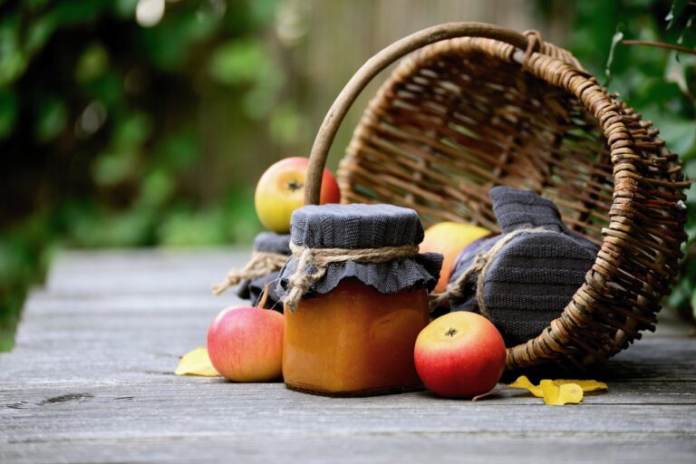 A glass jar of homemade preserve with a cloth lid tied with twine next to fresh apples and a tipped-over wicker basket on a wooden surface with a blurred green background.