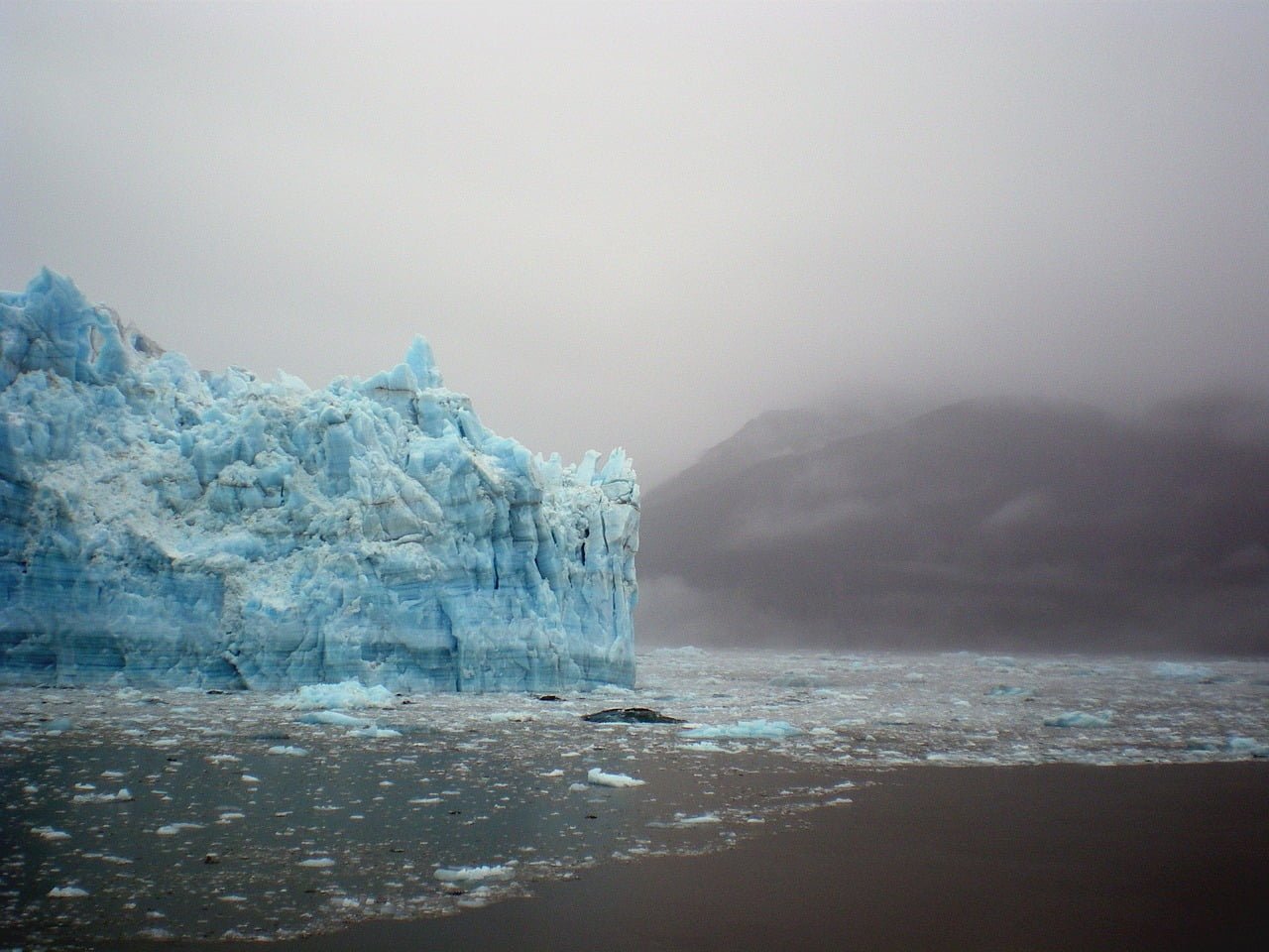 A towering blue glacier rising from a dark sea with scattered ice under a heavy, foggy sky, with obscured mountains in the background.