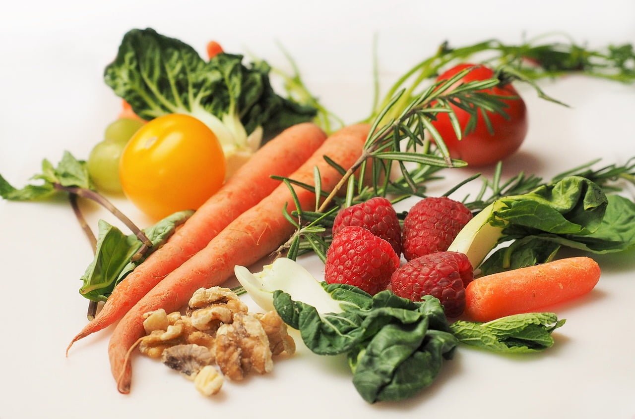 A variety of fresh vegetables and fruits on a white surface, including carrots, tomatoes, raspberries, grapes, spinach, a yellow cherry tomato, walnuts, and herbs.
