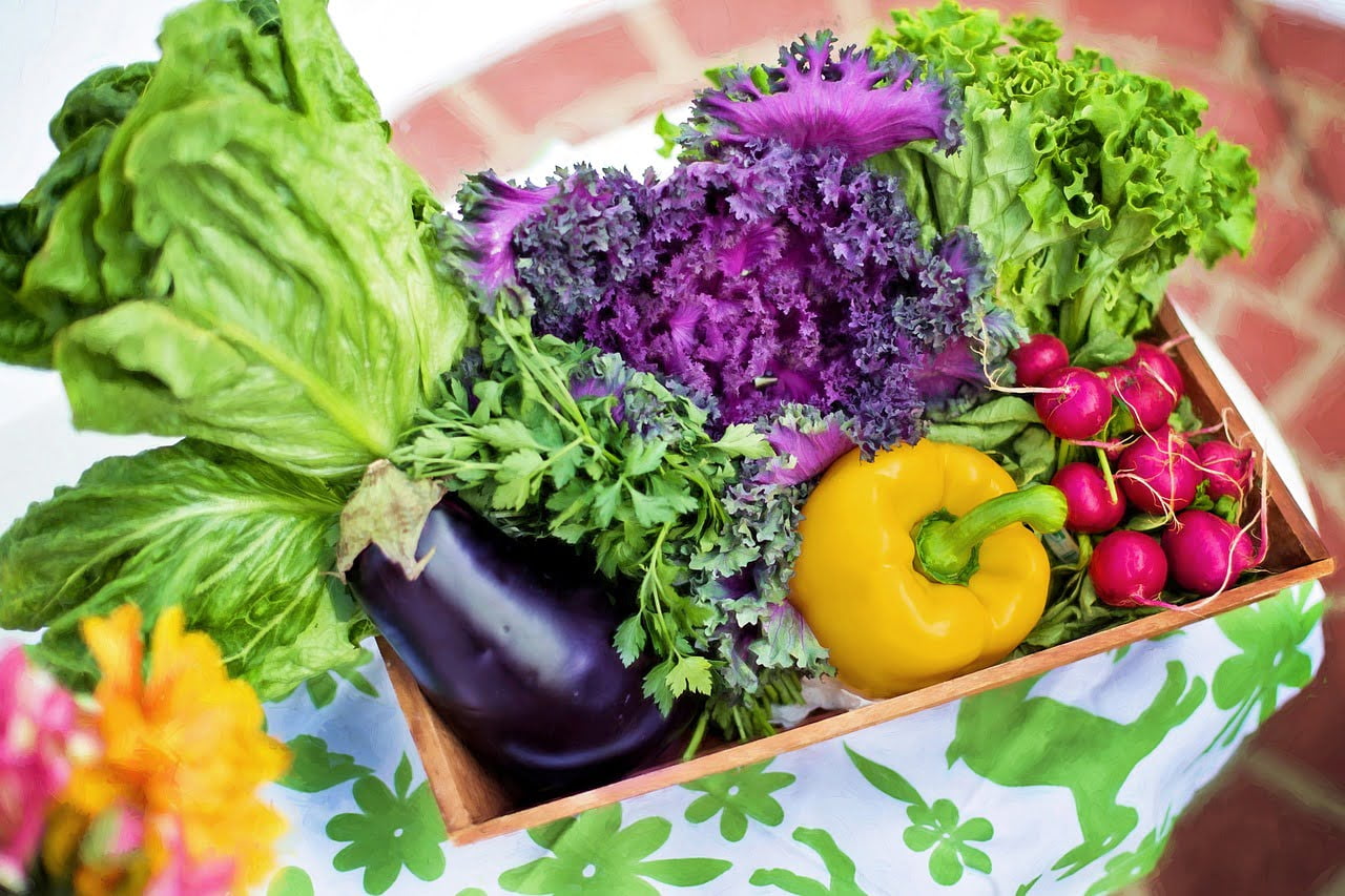 A wooden crate filled with an assortment of colorful fresh vegetables, including lettuce, purple kale, parsley, a yellow bell pepper, an eggplant, and radishes, placed on a table with a floral cloth.