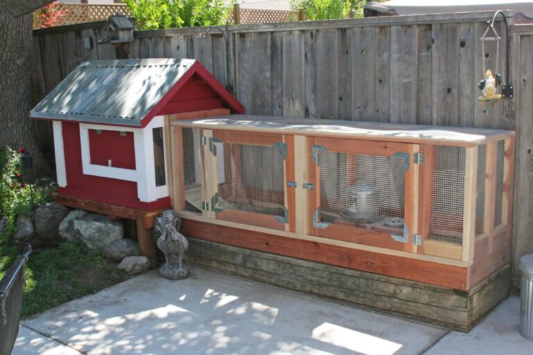 A homemade chicken coop with a red and white house connected to a fenced enclosure, placed against a wooden fence in a backyard.