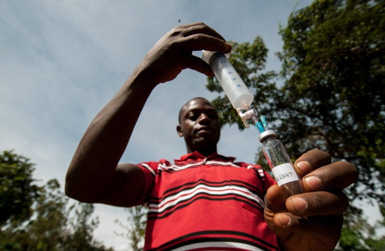 A person in a red striped shirt preparing a syringe with a vial, viewed from a low angle against a backdrop of trees and sky.