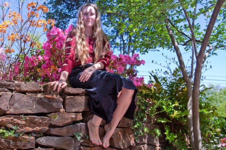 A woman sitting on a stone wall with colorful flowers in the background under a clear blue sky.