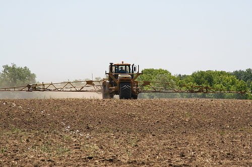 A tractor with an extended spray boom is treating a field, with trees in the background under a clear sky.