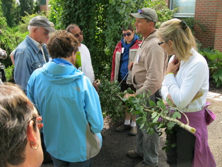 A group of people, some wearing jackets and caps, engaged in an outdoor gardening workshop or tour, with plants and a brick building in the background.