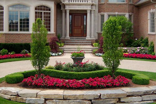 A symmetrical front yard landscaping of a large brick house with decorative shrubs, pink flowers, and a central stone planter.