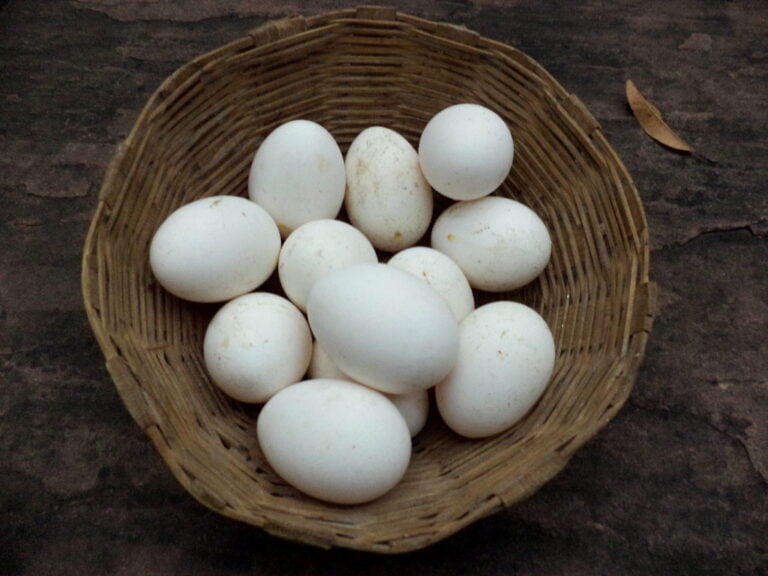 A wicker basket containing multiple white eggs on a textured surface with a dry leaf beside it.