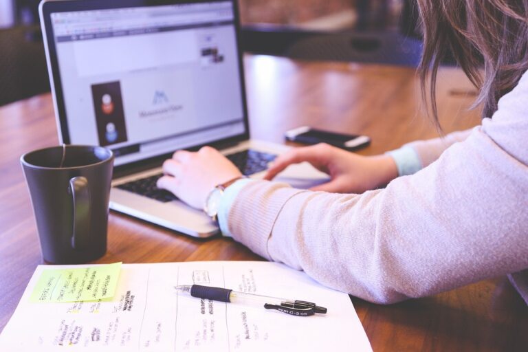 A person working on a laptop with notes and a pen in the foreground and a coffee mug beside the laptop.