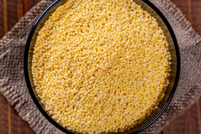 A close-up view of yellow millet grains in a black bowl on a burlap cloth with a wooden surface in the background.