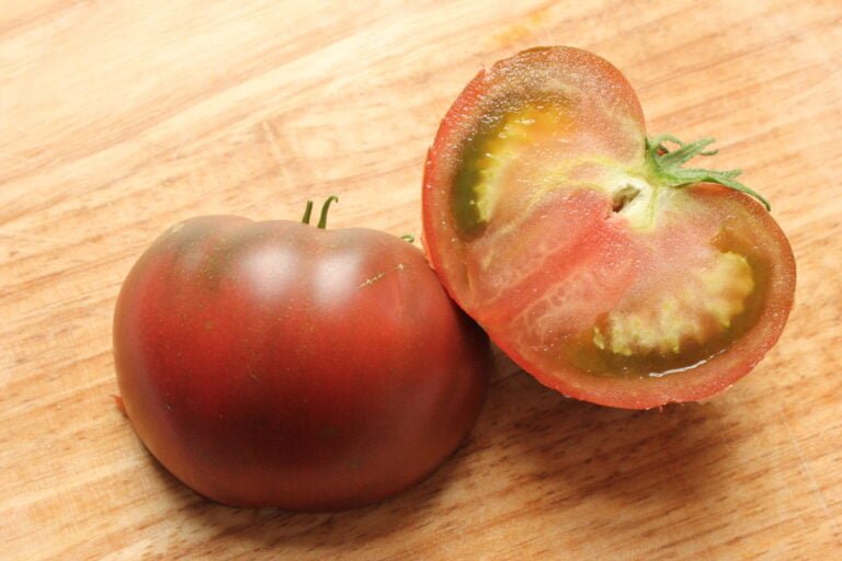 A whole and a halved tomato on a wooden cutting board. The halved tomato shows the seeds and juice inside.