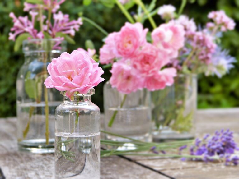 Several small glass vases with pink roses and purple flowers arranged on a wooden surface with a blurred green background.