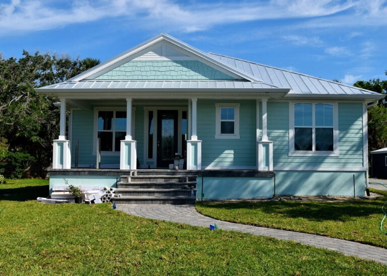 A teal single-story house with a front porch, white columns, a stone pathway, and a green lawn against a clear blue sky.