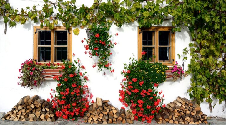 A white wall with three windows framed by wooden shutters, decorated with green vines and colorful flowers, with piles of chopped wood below.